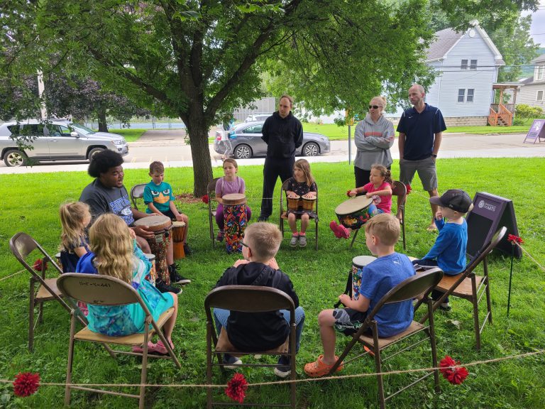 children with drums sit in circle of folding chairs with instructor