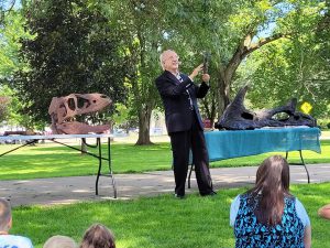 Mike the dinosaur hunter holds a claw for the audience with two skulls on tables in the background