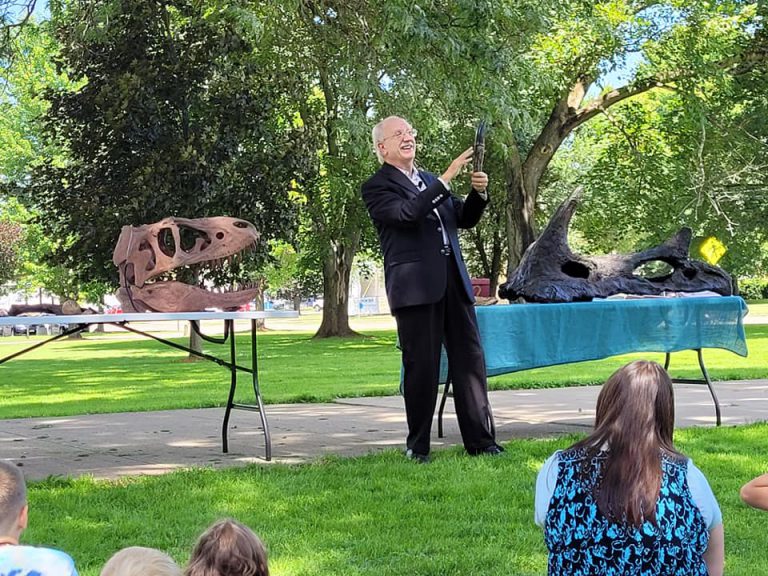 Mike the dinosaur hunter holds a claw for the audience with two skulls on tables in the background