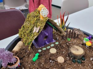 fairy garden with purple house with moss roof, wood slice path, tiny brown picket fence, and miniature plants and mushrooms