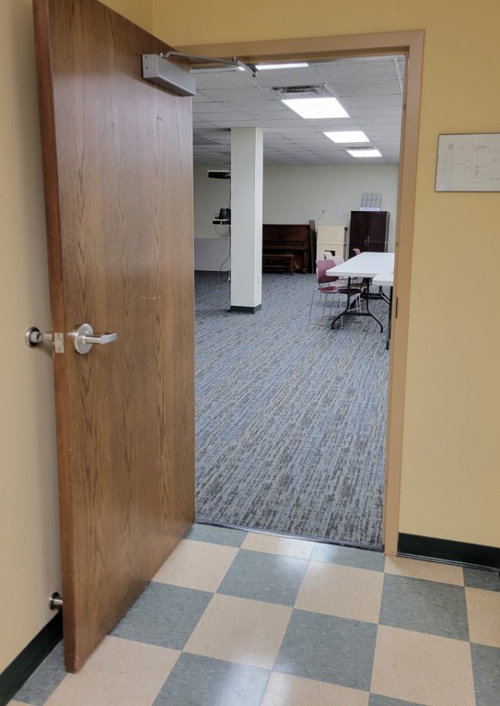 Open wooden door leading to a room with carpeted flooring and furniture.