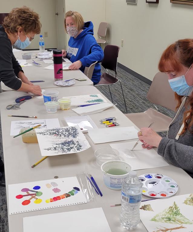 two women work on watercolor paintings as class instructor demonstrates technique