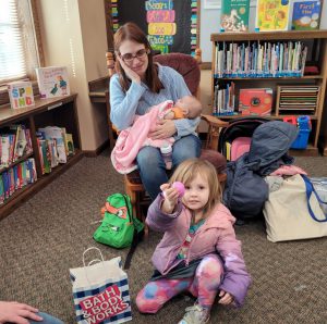 woman in rocking chair holding infant while young girl crouches with Easter egg