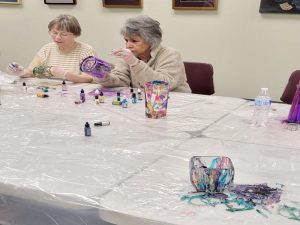 Two women add ink to their glassware; foreground: squat glass bowl or vase with several colors of ink