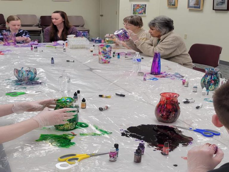 3 women and one girl work on glassware projects; foreground: glass vases with alcohol ink, one red and one green, with large puddles of ink on the table