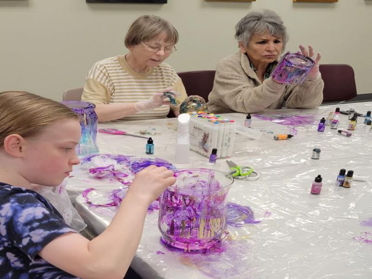 foreground: girl adds ink to her large wide vase of pink, purple and gold; background: two women add ink to their vases