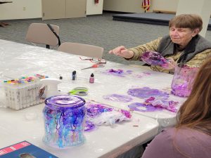 Woman reaches for ink bottle to add to her tall vase of pink and purple; foreground large wide glass vase with blue, purple and pink ink