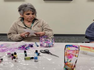 woman sits with wide glass vase decorated pink and teal inks. In foreground a tall glass vase decorated with a rainbow of ink colors