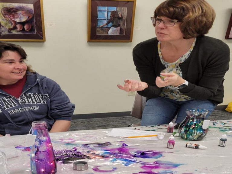 woman listens to fellow class participant as instructor Patience Wnek talks with another class participant. Glassware decorated with alcohol inks sits on the table.