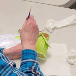 person holds paintbrush to use end of handle to paint white dots to decorate bright green mini birdhouse