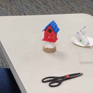 mini red birdhouse with blue stars and blue roof with white stars perched on mini white flower pot with red and white sprinkles and twine wrap around base.