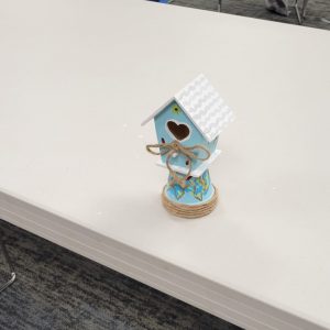 tiny light blue birdhouse with white and grey roof and heart cutout for door perched on light blue pot with blue flowers and green leaves