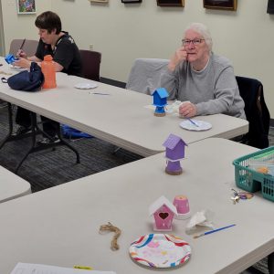 woman with dark blue mini birdhouse with light blue roof looks on as second woman paints decorative details on a second dark blue mini birdhouse