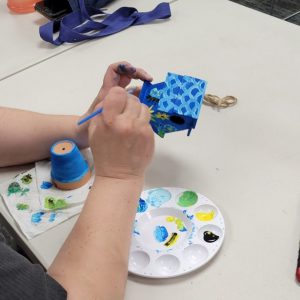 person paints dark blue mini birdhouse with light blue shingle details and yellow sunflowers