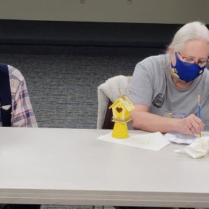 woman paints white decorative details onto a mini yellow birdhouse