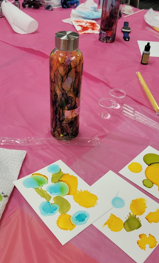 bottle with shades of red, purple, and green sits on a table next to papers with spots of green blue and yellow