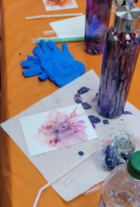 paper with pink and purple ink spots sits next to bottle painted with silver, purple blue and red. A bottle with purple and blue can be seen in the background.