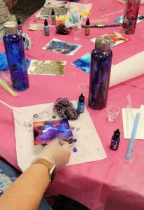 water bottle painted with blues and purples and bottle painted with purples and reds, surrounded by papers painted with spots of several colors of ink