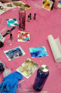 several papers with ink spots in shades of red, blue, purple, grey, yellow and green, with two water bottles, one in shades of red and purple and one in shades of purple and blue