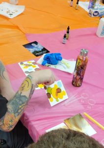 three papers with brown, red, green, yellow, and blue ink splotches sit near a bottle painted with drips in the same colors. A tattooed arm holds one of the papers up off the table