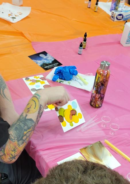 three papers with brown, red, green, yellow, and blue ink splotches sit near a bottle painted with drips in the same colors. A tattooed arm holds one of the papers up off the table