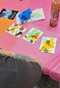 three papers with brown, red, green, yellow, and blue ink splotches sit near a bottle painted with drips in the same colors