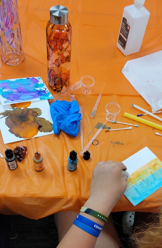 several papers with purple, blue, brown, and orange ink sit on a table next to a bottle with drips of red ink. Plastic cups, pipettes, and a bottle of rubbing alcohol are nearby