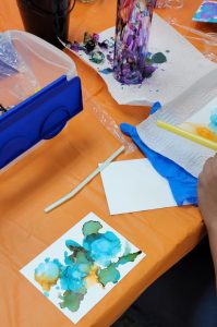 ink painting in green blue and orange sits on a table near paper towels and a bottle with ink drips in purple red and green