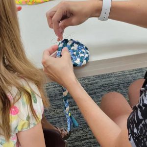 a pair of hands stitches a braided rope of fabric into a circular shape as a blond girl watches