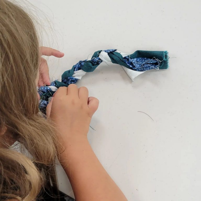 a young blond girl stitches a braided fabric rope into a circular rug