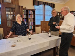 grey haired woman with glasses in dark blouse stands at a table with bottles of wine. Two men stand nearby, one with greying red hair and glasses and one with grey hair glasses and a beard