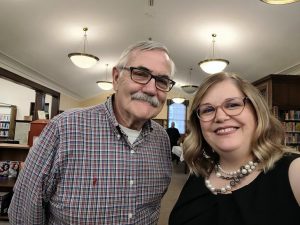 grey haired man with mustache and glasses wearing plaid shirt stands with blond woman with glasses in a black dress