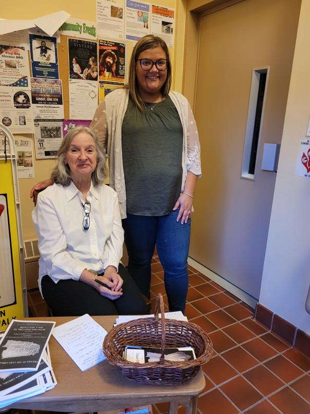 grey haired woman in white button down sits next to brown haired woman with glasses in white open shirt with olive tshirt and blue jeans next to small table with basket and pamphlets