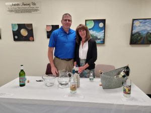 man with shaved hair and glasses in blue polo and khakis smiles with arm around brown haired woman in a white shirt with black cardigan and grey patterned skirt