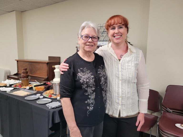 woman in black cardigan with short grey hair and glasses stands with red haired woman in white button down