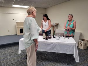 woman with long black hair and woman with short grey hair stand behind table with several bottles of wine, a third woman with greying blond hair talks to them