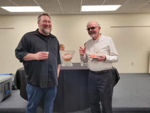 tall bearded blond man in black button down shirt and jeans stands with grey haired man with glasses in white button down and black slacks