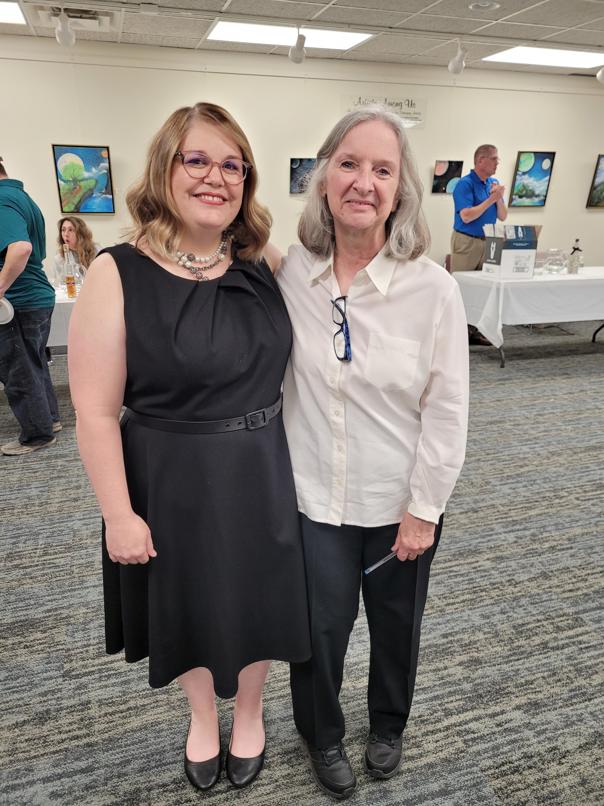 blond woman with glasses in a black dress stands with grey haired woman in black pants and white button down shirt