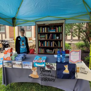 woman in Cuba Library t-shirt stands behind table with grey Cuba Library tablecloth with postcards, covid test kits, sanitizer, wipes, and library t-shirts tote bags and water bottles