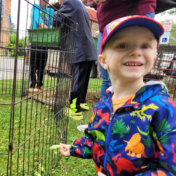 young blond haired boy with a hat and dinosaur patterned coat smiles toward the camera next to an animal enclosure