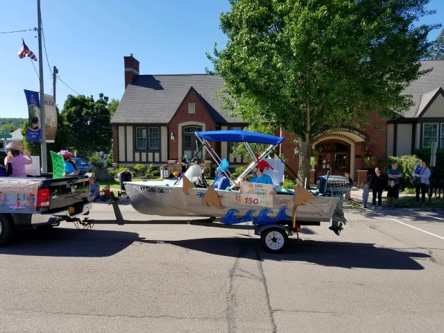 small silver fishing boat with blue roof with cardboard waves attached being towed by black pickup. people with silly fish hats ride in the boat and truck