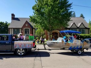 small silver fishing boat with blue roof with cardboard waves attached being towed by black pickup. people with silly fish hats ride in the boat and truck