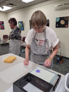 woman with short brown hair bends over table with sheet of white handmade paper with purple star