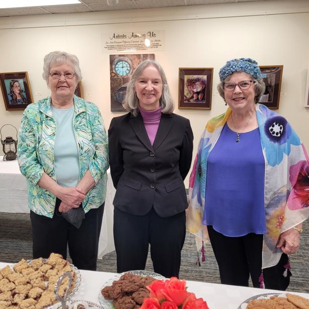 a trio of ladies smile for the camera with a table of cookies in front of them