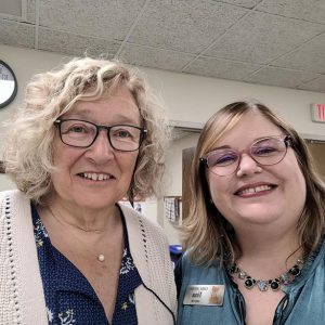 woman with graying curly blond hair and glasses smiles with woman with straight blond hair and glasses