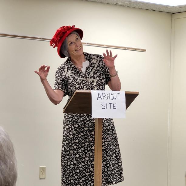 woman in a black flowered dress and red hat stands at lectern labeled "Arnout Site"