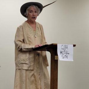 woman in a tan dress with matching jacket and brown hat with long feather stands at a lectern labeled "Cuba Hotel Site"