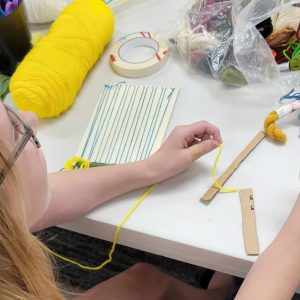 girl holds strand of yellow yarn, a weaving board sits under her wrist.