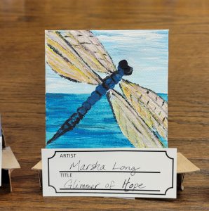 painting of light blue sky, blue water and a blue dragonfly with pink and yellow wings