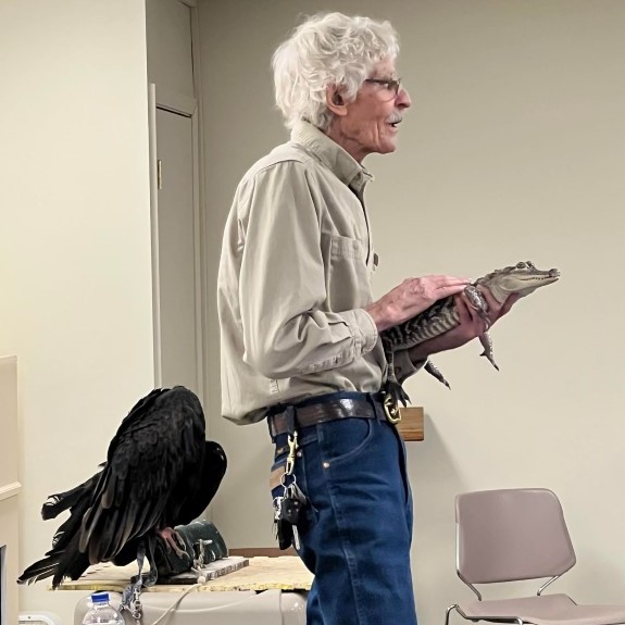 slim man in tan shirt and blue jeans with white hair holds caiman while vulture perches on table behind him